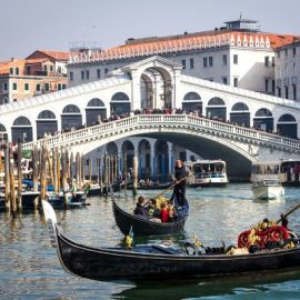 Rialto Bridge Italy with 2 gondolas on the canal with people inside. There are buildings in the background.