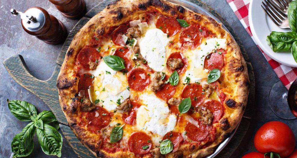 A pepperoni pizza with basil leaves on top on a pizza board. There are tomatoes, basil, a glass of wine, salt and pepper mills and a plate with forks on a red and white checkered napkin. This is for a blog about national pizza day. Photo by Canva.